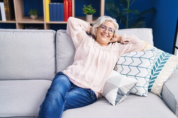 Wall Mural - Middle age grey-haired woman relaxed with hands on head sitting on sofa at home