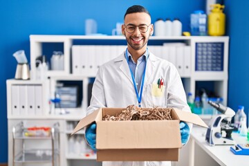 Poster - Young hispanic man working at scientist laboratory holding cardboard box winking looking at the camera with sexy expression, cheerful and happy face.