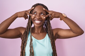 Canvas Print - African american woman standing over pink background doing peace symbol with fingers over face, smiling cheerful showing victory