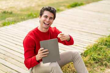 Wall Mural - Young handsome man holding a tablet at outdoors points finger at you with a confident expression