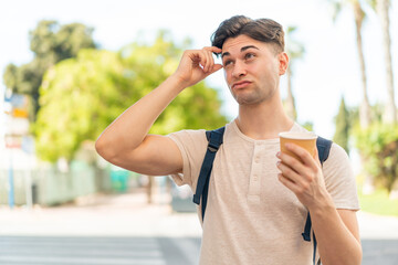 Young handsome man holding a take away coffee at outdoors having doubts and with confuse face expression