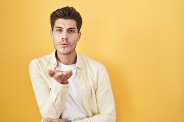 Canvas Print - Young hispanic man standing over yellow background looking at the camera blowing a kiss with hand on air being lovely and sexy. love expression.