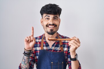 Wall Mural - Young hispanic man with beard wearing apron tasting food holding wooden spoon surprised with an idea or question pointing finger with happy face, number one