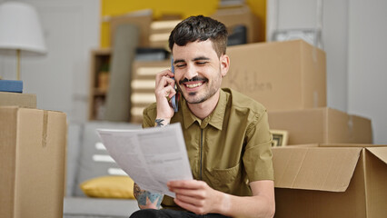 Canvas Print - Young hispanic man talking on smartphone reading document at new home