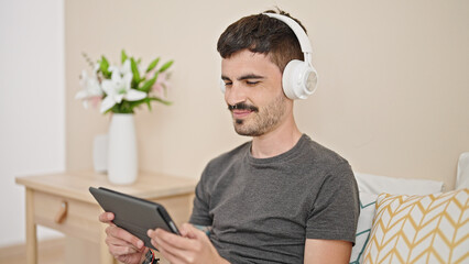 Wall Mural - Young hispanic man using touchpad and headphones sitting on bed at bedroom