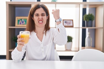 Wall Mural - Brunette woman drinking glass of orange juice angry and mad raising fist frustrated and furious while shouting with anger. rage and aggressive concept.