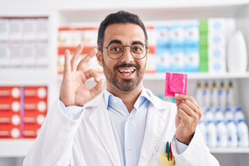 Wall Mural - Hispanic man with beard working at pharmacy drugstore holding condom doing ok sign with fingers, smiling friendly gesturing excellent symbol