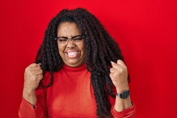 Poster - Plus size hispanic woman standing over red background very happy and excited doing winner gesture with arms raised, smiling and screaming for success. celebration concept.