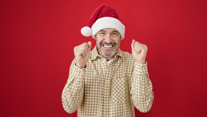 Wall Mural - Middle age man smiling confident wearing christmas hat over isolated red background