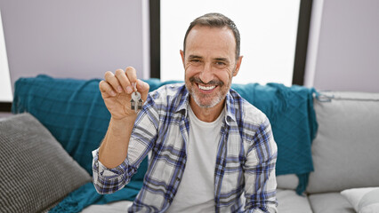 Wall Mural - Smiling grey-haired middle-aged man, confidently holding key while sitting on a sofa, radiating positivity in his new home, enjoying the happiness of a successful real estate investment.