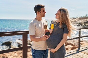 Canvas Print - Man and woman couple expecting baby using smartphone at seaside