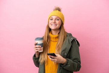 Wall Mural - Young blonde woman wearing winter jacket isolated on pink background holding coffee to take away and a mobile