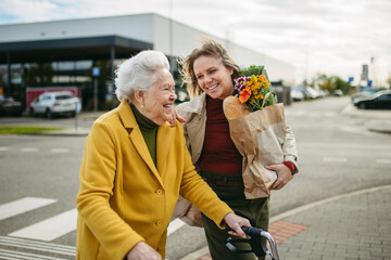 Mature granddaughter carrying grandmother's shopping bag. Senior woman and caregiver going to home with groceries from supermarket, during cold autumn day.