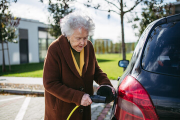 Close up of beautiful senior woman plugging charger in her electric car. Progressive elderly woman charging her electric vehicle on street.