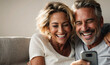 © anandart - Young mature couple laughing looking at mobile phone sitting on sofa at home, happy diverse husband and wife using online services on internet, technology lifestyle concept, space for text