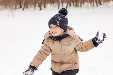 Little boy having fun playing with fresh snow during snowfall. Snowball fight. Kid dressed in warm clothes, hat, hand gloves and scarf. Active outdoors leisure for child on nature in snowy winter day.