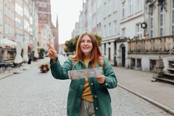 Wall Mural - Attractive young female tourist is exploring city. Redhead girl with backpack pointing finger and holding a paper map on city street in Gdansk. Traveling Europe in autumn.