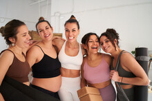 Group of beautiful sporty women looking at camera. Smiling happily together in yoga class. Cheerful fitness friends hugging in gym. Young female, healthy lifestyle in community and friendship