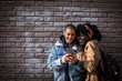 © Marko Geber - Couple of gay women using smartphone together leaning on brick wall