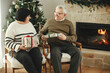 © sonyachny - Beautiful elderly couple exchanging christmas gifts and sitting at warm fireplace in stylish festive decorated living room. Happy senior family enjoying cozy evening with presents. Happy holidays