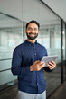 © insta_photos - Happy professional Indian businessman using tab computer working standing in office. Smiling professional business man employee holding digital tablet tech device working, looking at camera. Vertical