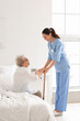 © Pixel-Shot - Young caregiver helping senior woman with stick to stand up in bedroom