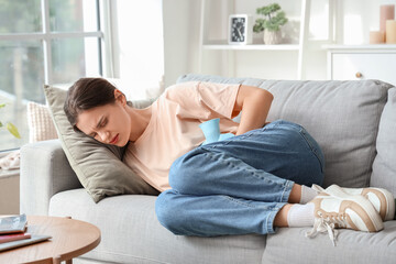 Sticker - Young woman with hot water bottle lying on sofa and suffering from abdominal pain at home