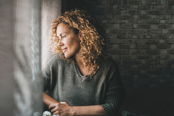 Wall Mural - One serene adult woman looking outside the window at home with peaceful expression on face and little smile. Confident female people in indoor relax leisure activity alone. Dreaming and thinking lady