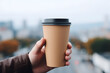 Man's hand holding brown disposable coffee cup with blurry city in background
