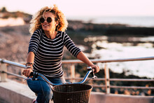 Cheerful And Happy Adult Young Woman Enjoy A Bike And Use Green Alternative Environment Way Of Transport With Ocean Beach Coast In Background. Active Leisure People Using Bicycle Outdoor