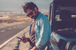 © simona - Traveler man use phone mobile cnnection standing against a black off road car with long road in background. Road trip and travel lifestyle for adventurer male people. Use of digital map road trip