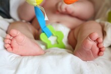 The Baby Is Sitting In A Child's Chair. A Baby In A Nappy Holds A Rattle In His Hands. Feet In The Foreground