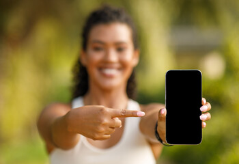 fitness woman in sportswear pointing at blank phone screen outdoors