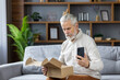 © Tetiana - Shocked senior gray-haired man sits on the sofa at home, holds the phone in his hands and looks sadly at the open package he received in the mail
