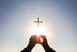 Fototapeta  - Brightly shining sunlight and silhouette of Christian hands holding high the cross of Jesus Christ symbolizing death and resurrection
