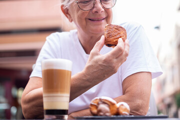 Sticker - Senior woman sitting at cafe table in the morning having breakfast with sweet food and a glass cup filled with coffee and milk.