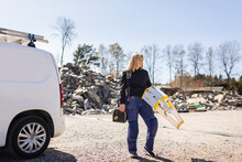 Smiling Female Electrician Carrying Ladder And Toolbox Standing On Road By Van