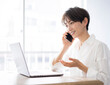 © lielos - 自宅でリモートワークをしている女性。/彼女はノートパソコンの画面を見ながらスマートフォンで話ている。