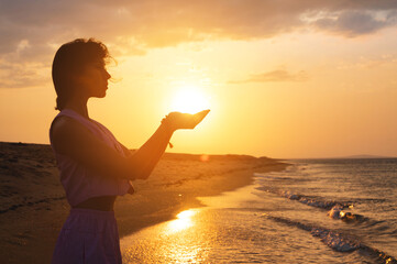 Wall Mural - Portrait of a young woman in the form of a silhouette and hands holding the sun. Sunset over the sea, the sun on the palms of a calm girl