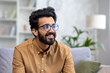 © Liubomir - Close-up photo of a young Indian man in glasses sitting on the couch at home and looking to the side with a smile.