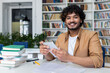 © Liubomir - Portrait of a young Indian male student sitting in the university library, holding a tablet and smiling at the camera.