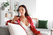 Beautiful young Asian woman with glass of wine sitting on sofa in living room