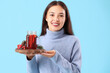 Pretty young woman holding tray with glass of warm mulled wine and Christmas decor on blue background