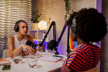 Wall Mural - Serious multiracial girls are sitting in a home recording studio and filming a podcast while wearing headphones and discussing topics.