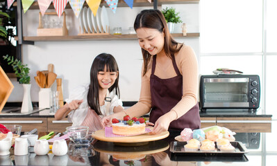 Asian beautiful female baker pastry chef mother wears apron standing smiling helping teaching little pretty girl daughter decorating cake with mixed berry fruit making homemade bakery in home kitchen