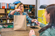 © David Pereiras - Smiling blonde young woman shop assistant delivering kraft paper bag with purchased clothing items to female customer. Happy woman employee taking credit card of client to charge purchase in store.