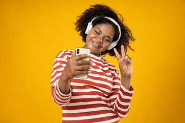 Wall Mural - Curly young woman with headphone and smartphone on yellow background
