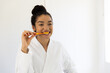 © WavebreakMediaMicro - Happy biracial woman in bathrobe brushing teeth in sunny bathroom
