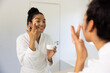 Happy biracial woman in bathrobe applying cream on her face in sunny bathroom