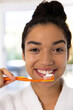 © wavebreak3 - Portrait of happy biracial woman in bathrobe holding brush with tooth paste in sunny bathroom